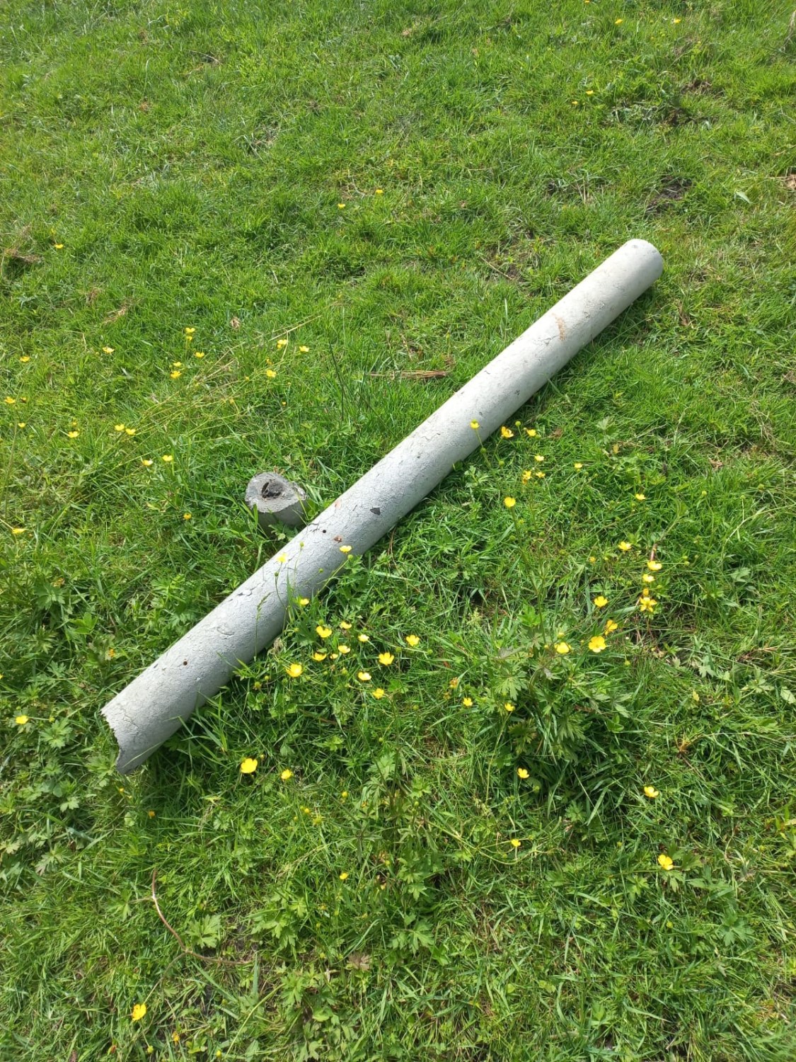 Ein gebrochener grauer Rohrabschnitt liegt auf grünem Gras mit gelben Blumen.